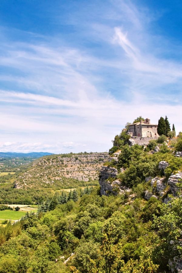 An old stone chapel, a church or a monastery standing on the edge of a rock. Beautiful view from the mountain to the valley of the river with fields, meadows, roads, white fluffy clouds and blue sky.